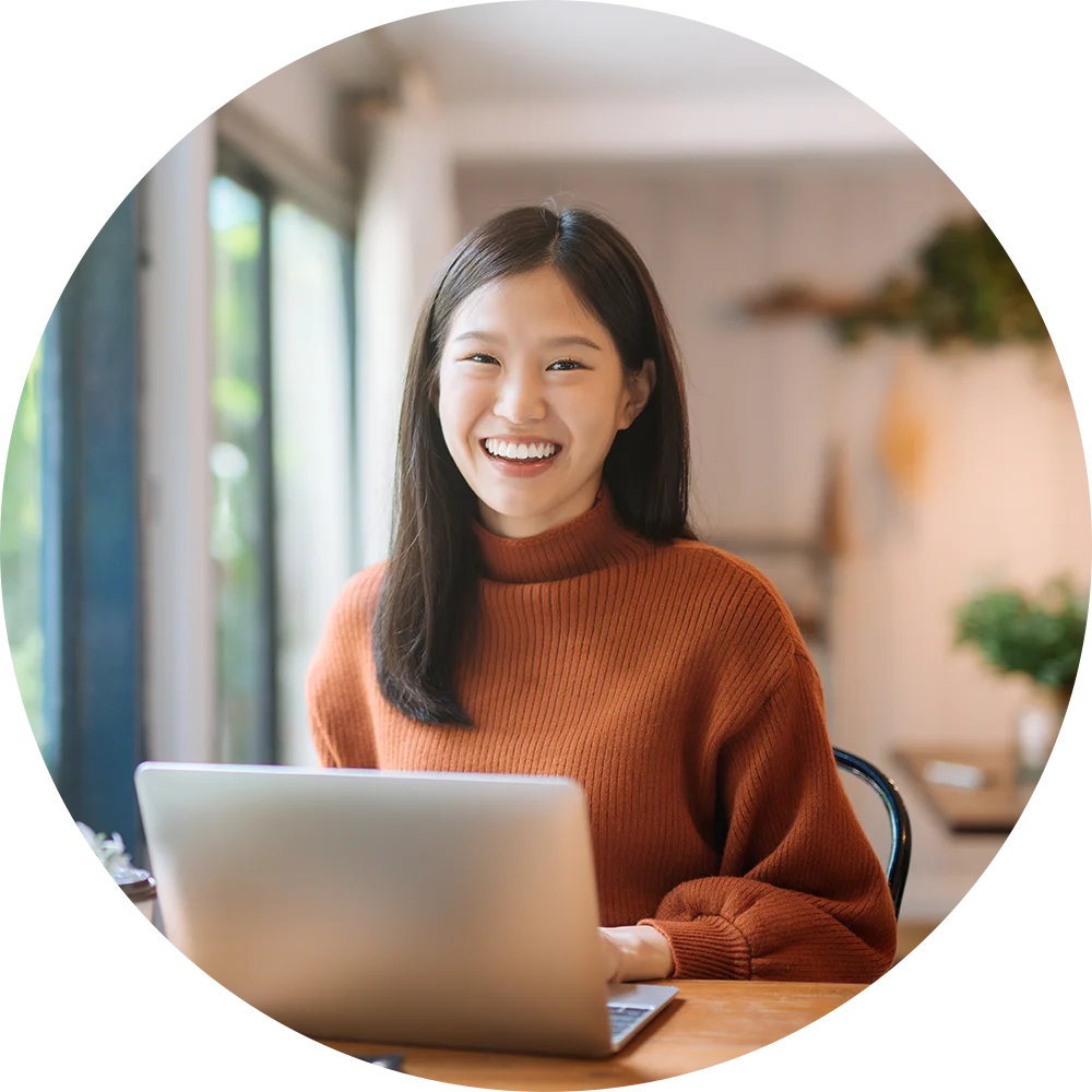 A smiling young woman uses a laptop at a table. She enjoys work and life after addiction treatment at Juniper Grove Recovery.