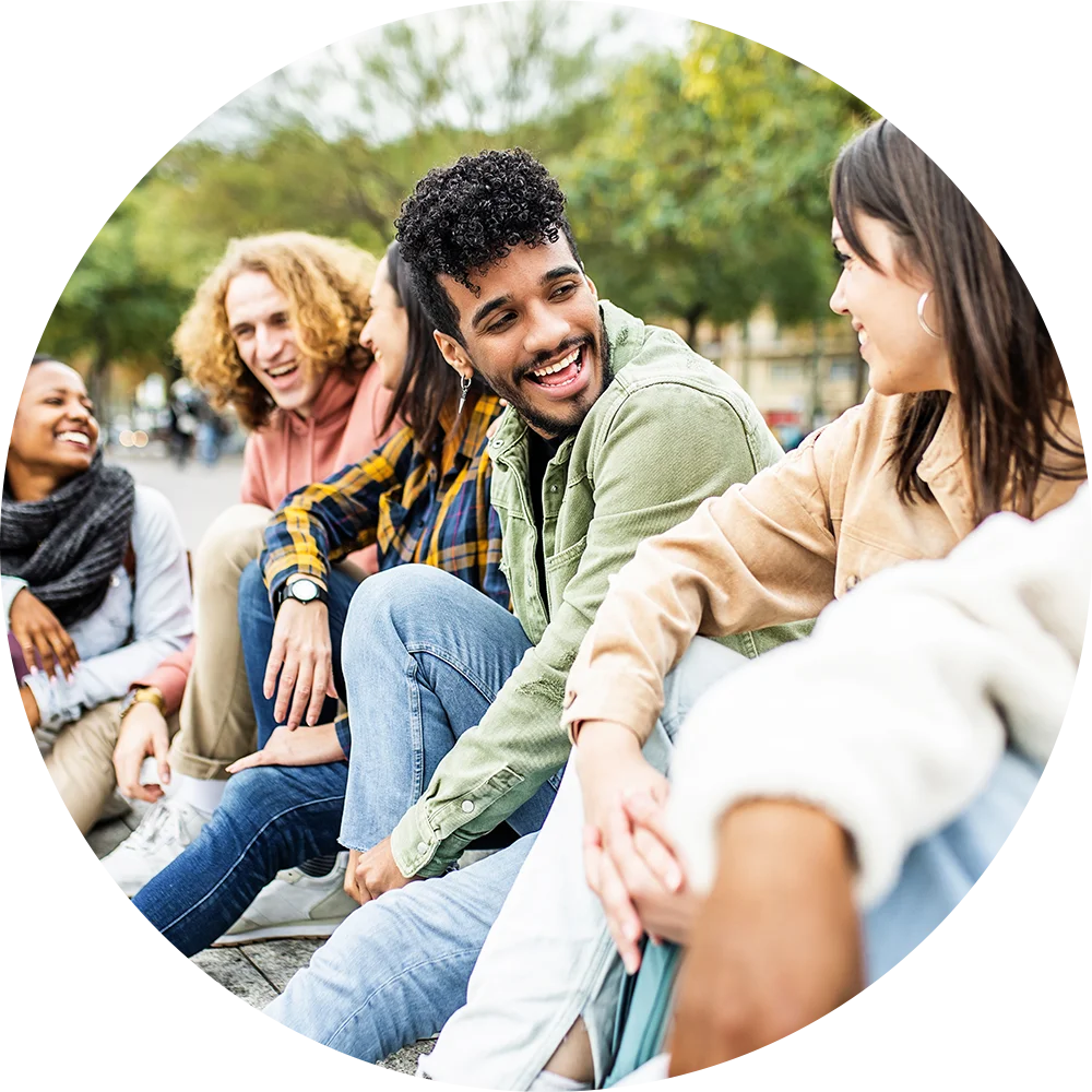 A group of young people, both male and female, are sitting outside. They're smiling, laughing, and enjoying life after addiction treatment at Juniper Grove Recovery.