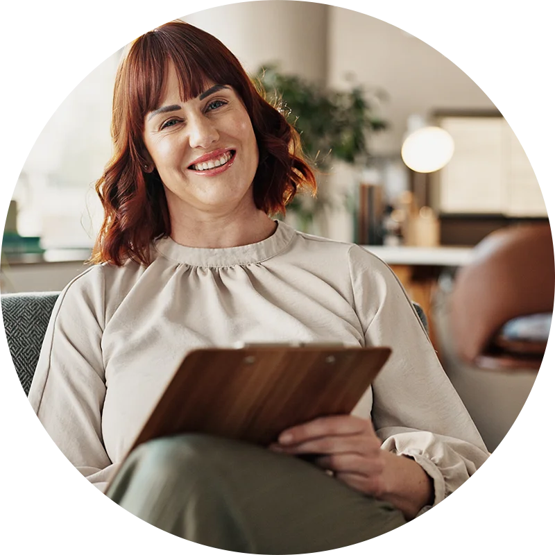 A therapist holding a clipboard, leans back and smiles at her client during an addiction therapy session at Juniper Grove Recovery.